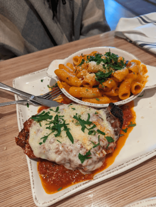 A plate featuring chicken topped with melted cheese and herbs, alongside a bowl of pasta in a creamy sauce.